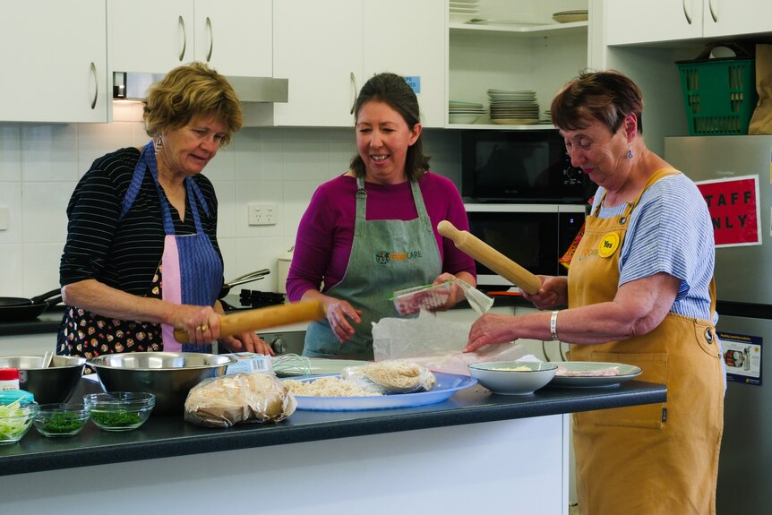 Three women in aprons stand behind a kitchen bench. Two are holding rolling pins.