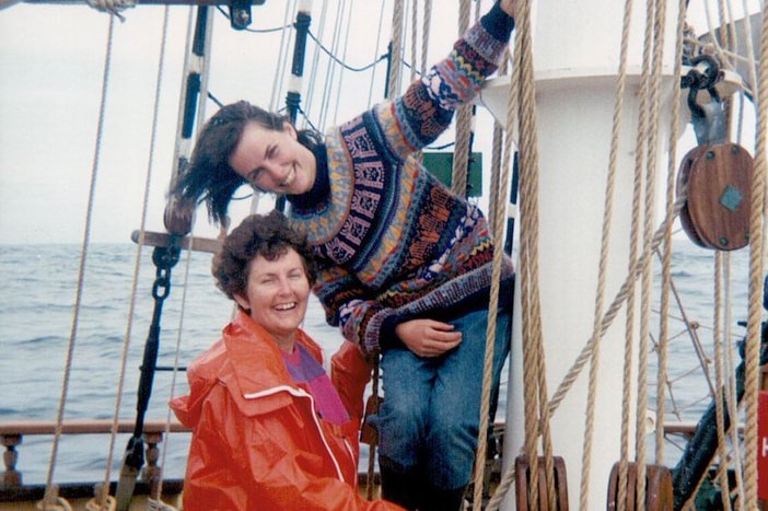 A mother and daughter hang onto the rigging of a sailing ship and laugh