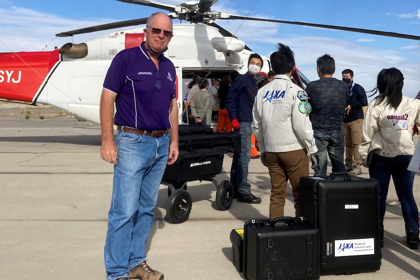 A man in a purple polo shirt stands in front of a helicopter and another group of people carrying a black box