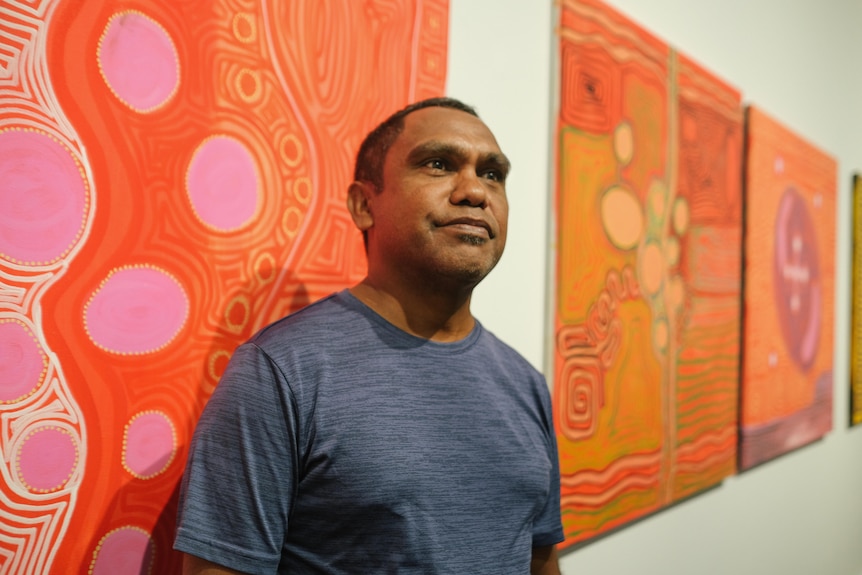 Senior Walmajarri artist Murungkurr Terry Murray stands in front of his vibrant art works, in a gallery