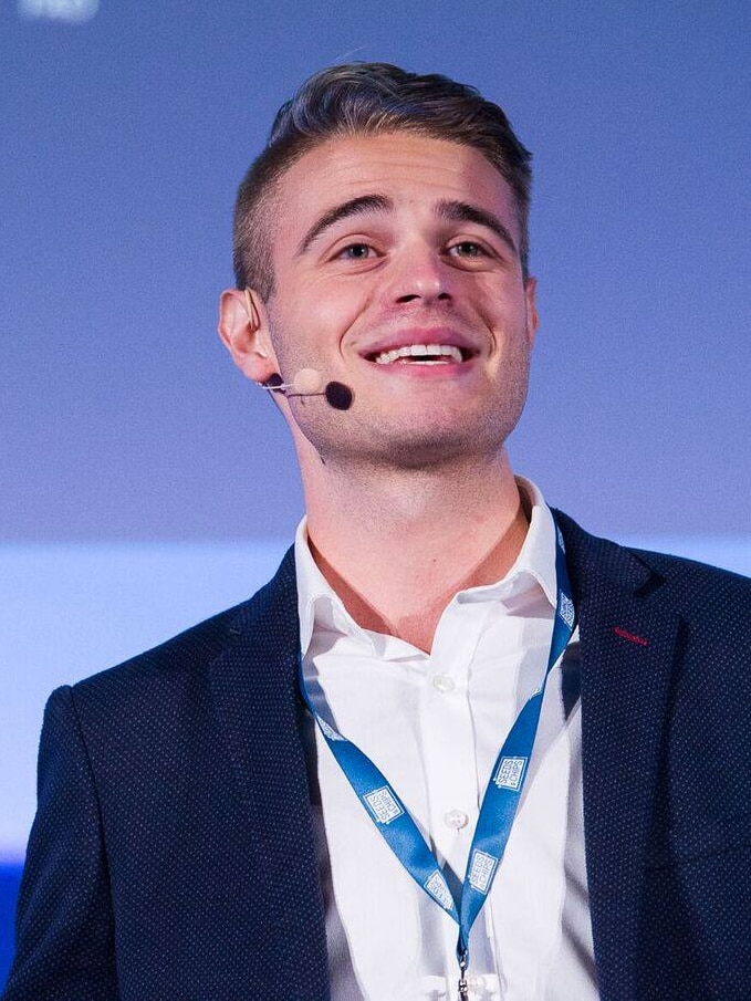 A mid shot of a young man in a suit looking up with a microphone headset and blue background. 