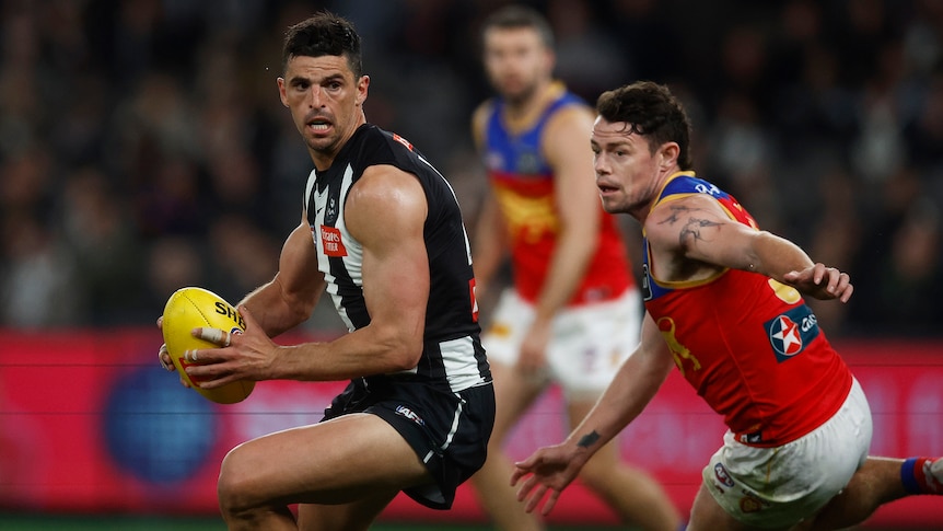A Collingwood AFL player looks behind him as he carries the ball, while a Brisbane player pivots to try and close in.