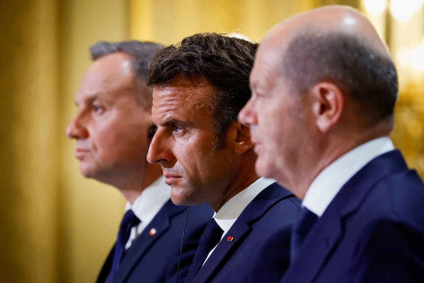 Emmanuel Macron, Olaf Scholz, and Andrzej Duda stand in a line wearing similar navy suits while looking forward.