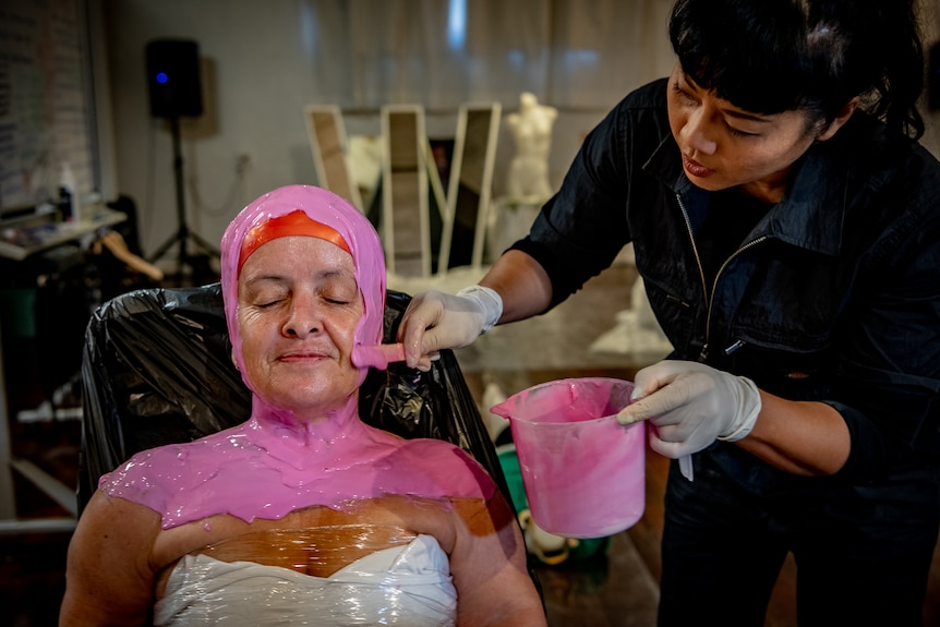 Leisa Prowd sits back in a chair and smiles while an artist applies pink moulding plaster to her face.