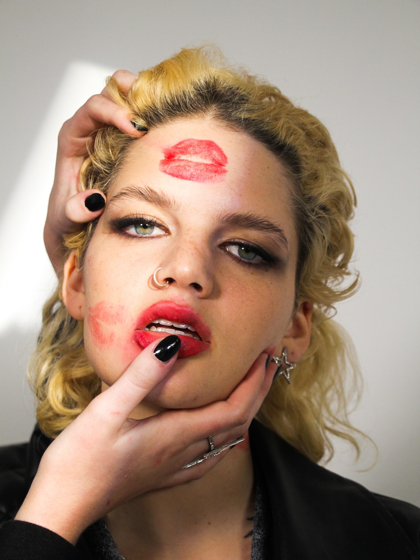 White blonde woman with two red lipstick marks on her face, and two hands clasping the bottom and side of her head