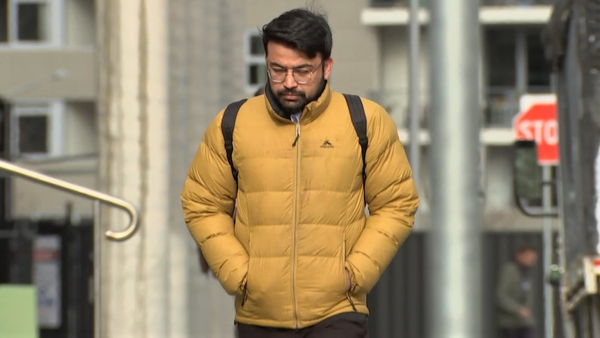 A man in a yellow puffer jacket, walking with his hands in his pockets and his head down.