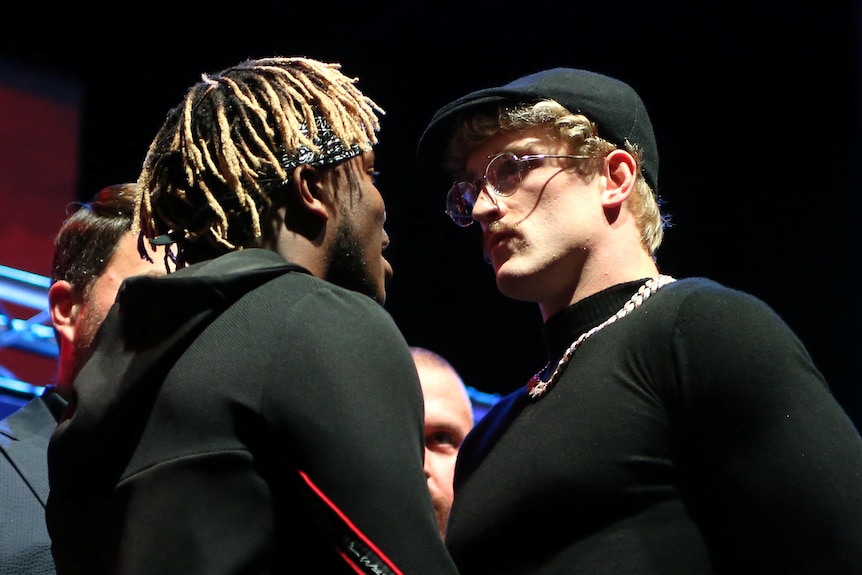 Mid shot of KSI (left, a black man with dreads & back to camera) faces a pale, stern Logan Paul with curly blond hair & dark cap