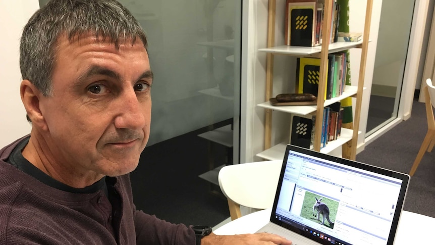 A man with dark brown and grey hair, wearing a maroon top, sits in front of a computer.