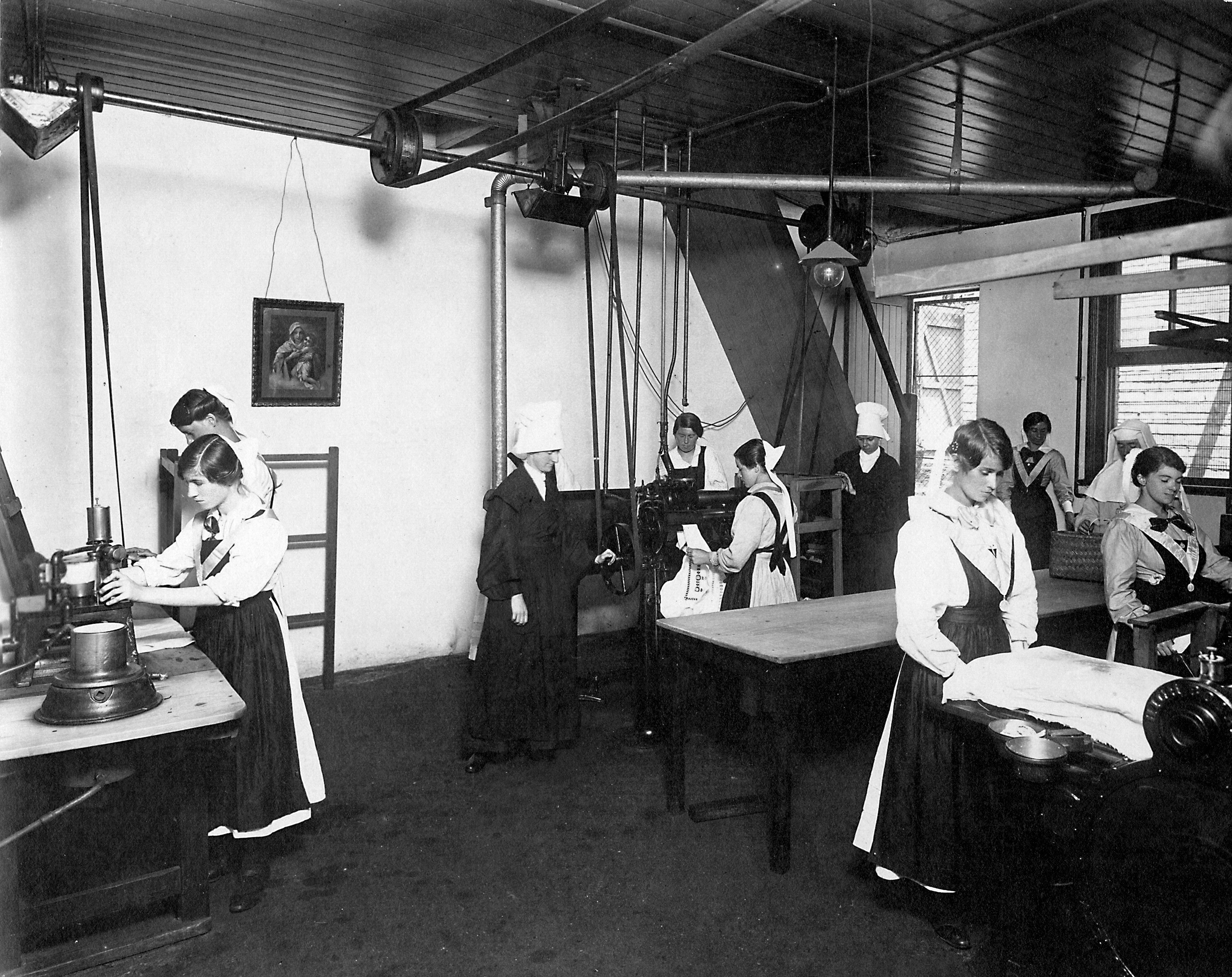 Black and white grainy photo of women in long white dresses and aprons folding sheets in a large room.