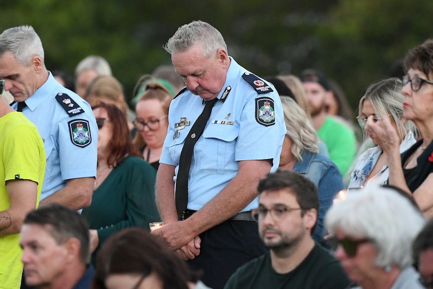 A police officer stands with arms folded head down, among a crowd.