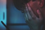 A close up image of a male teen with his hand over his face looking at a mobile phone
