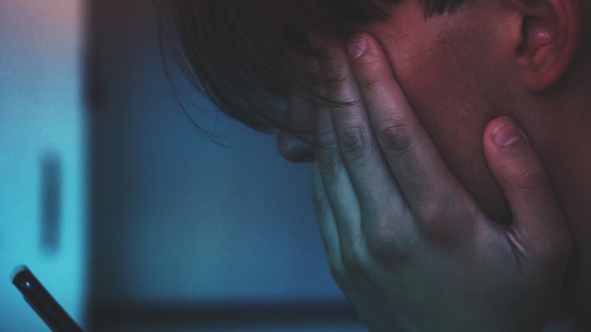 A boy holds his face in his hands and looks at a mobile phone.