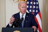 President Joe Biden raises a finger as he speaks from beind a podium in front of an American flag.