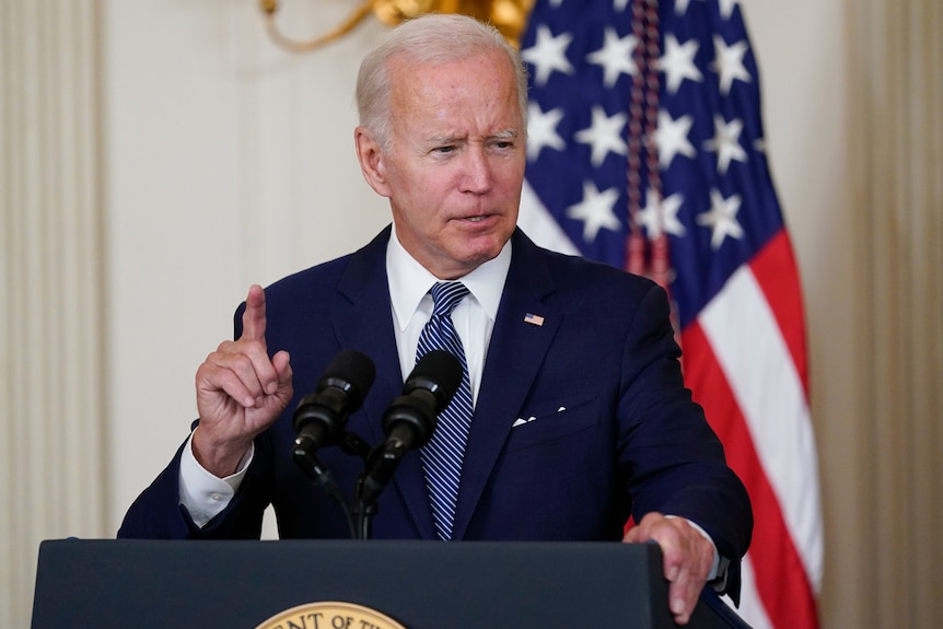Președintele Joe Biden ridică un deget în timp ce vorbește de pe podiumul din spate, în fața drapelului american.