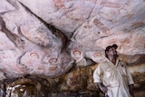 An Indigenous man in a khaki shirt and hat stands in a cave marked with rock art on Wunambal Gaambera country in WA's Kimberley.