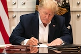 Wearing a suit and jacket, Donald Trump writes something on a piece of paper. The American flag is behind him