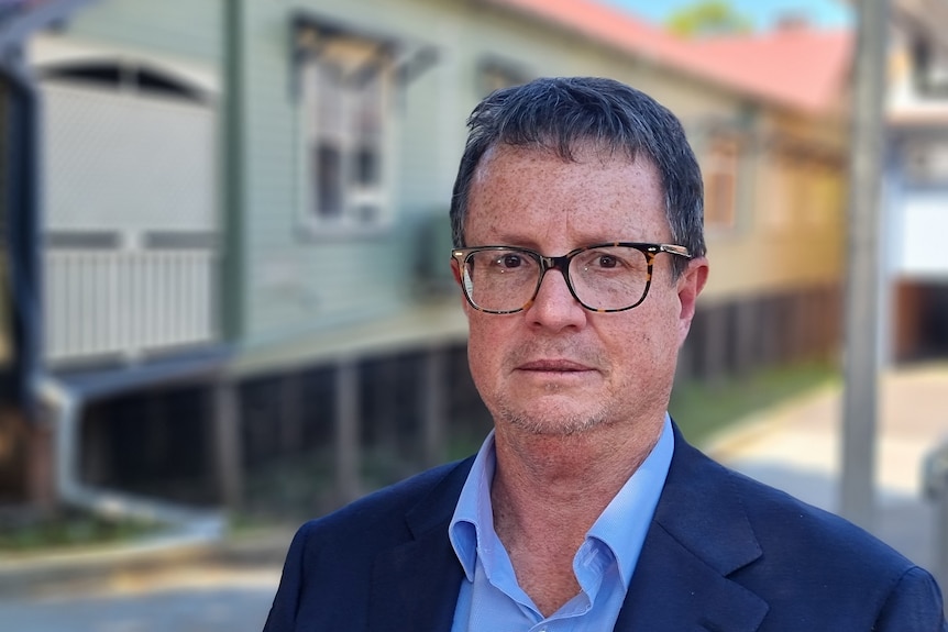 A man in glasses looks at the camera in front of a building