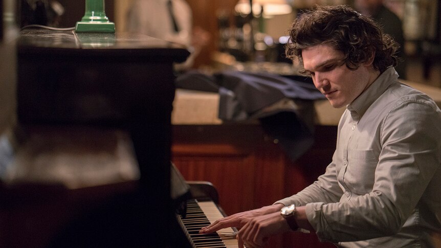 A man wearing light coloured button up shirt looks down as he plays an upright piano inside a room.