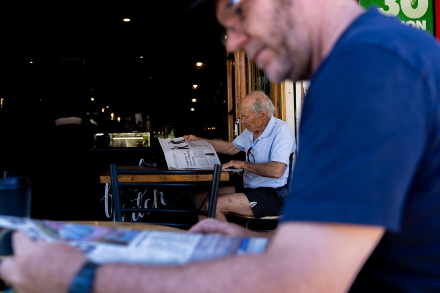 Two men, one in the foreground and one in the background, sit at a cafe and read a newspaper.