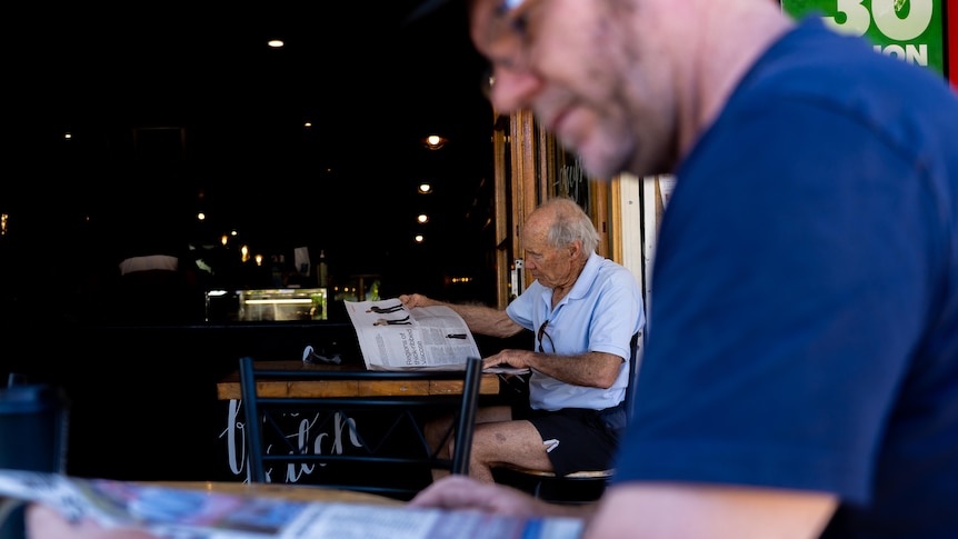 Two men, one in the foreground and one in the background, sit at a cafe and read a newspaper.
