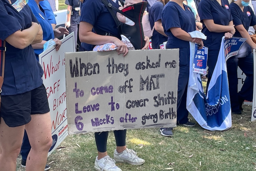 Crowd of people, below shoulders visible, woman holding cardboard  sign