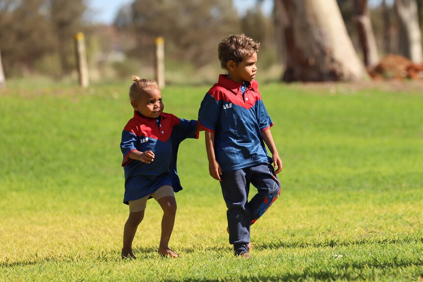 A young Aboriginal boy and girl wearing football colours stand on green football ground