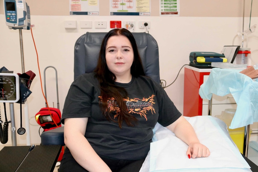 Tara Harrison sits in hospital chair with medical equipment around her, looking at camera