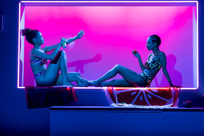 Two women in bikinis apply suntan lotion and chat. They're on stage, and the lights are hot pink.