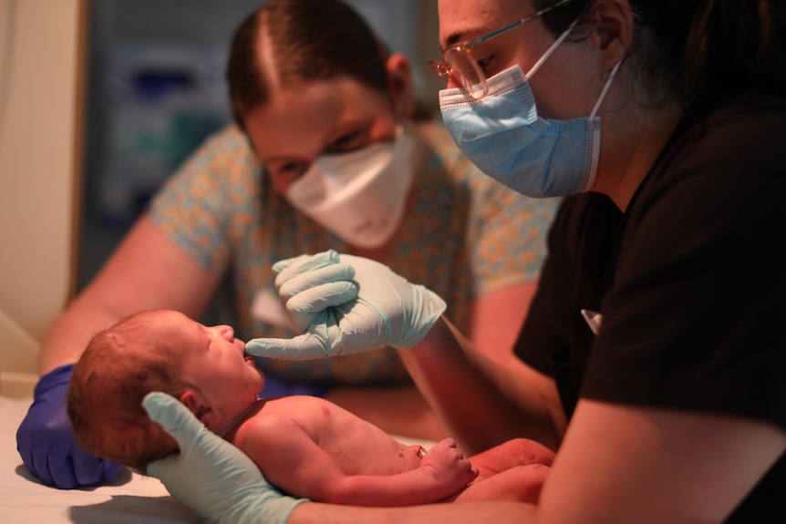 Midwife Bailey and student midwife Bella dote on a newborn, touching its mouth with a gloved finger.
