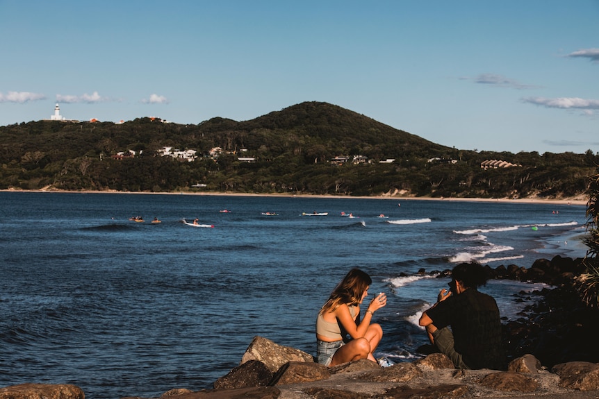 Two people sit on the rocks outlooking the blue water of Byron Bay's coastline