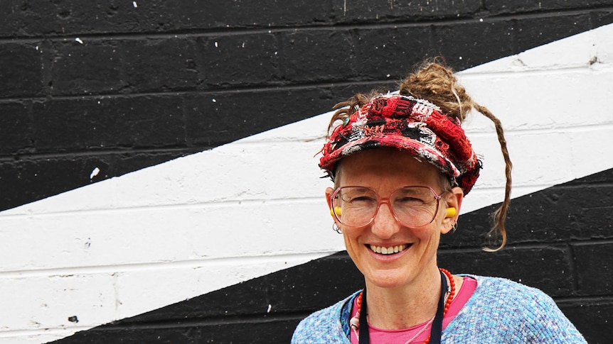 A woman poses in front of a striped wall in colourful clothing, big glasses and earplugs.
