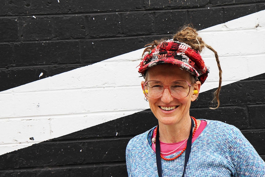 A woman poses in front of a striped wall in colourful clothing, big glasses and earplugs.