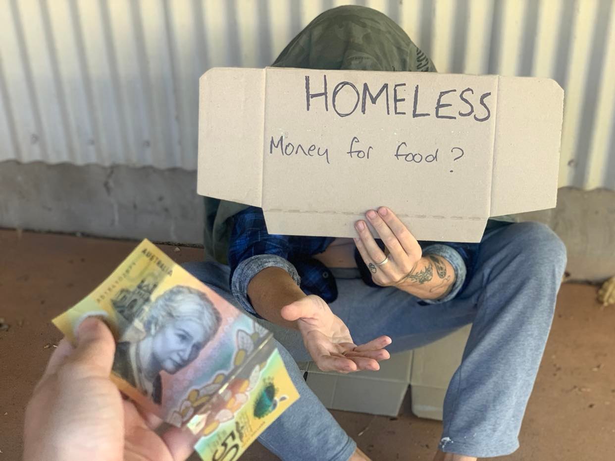 A person hands a $50 note to a man sitting on the ground holding a 'homeless' sign