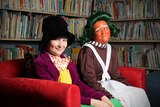 Children dressed as Willy Wonka and an oompa loompa sit on a couch in front of a book case.