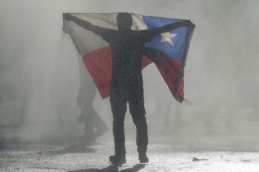 An anti-government protester stands in a cloud of tear gas holding up the red white and blue Chilean flag