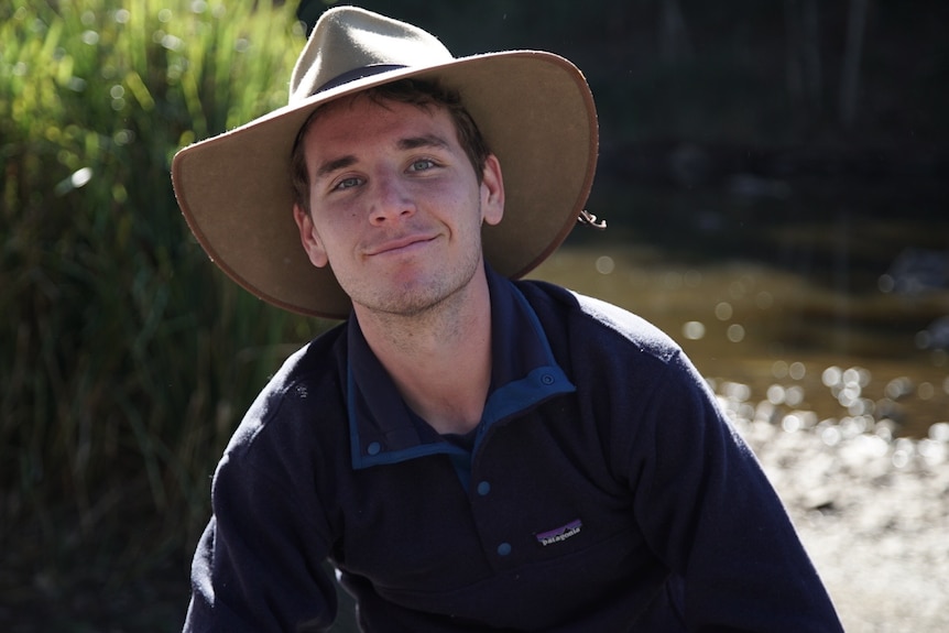 Zach Robba wearing a wide brim hat, smiling, long sleeve shirt, water and tall grass behind.