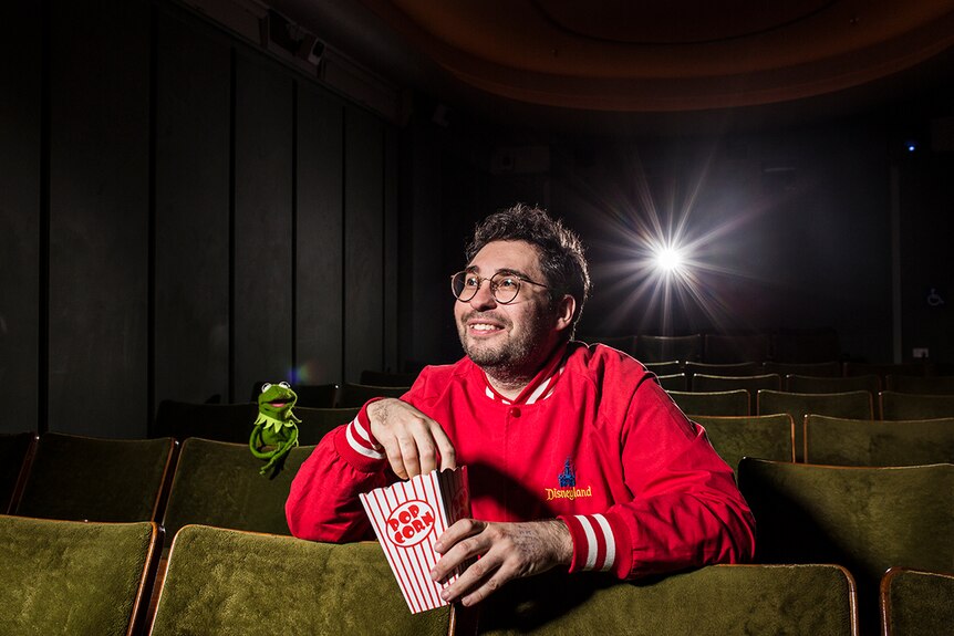 Colour photograph of artist David Capra eating popcorn in a cinema with Kermit the frog.