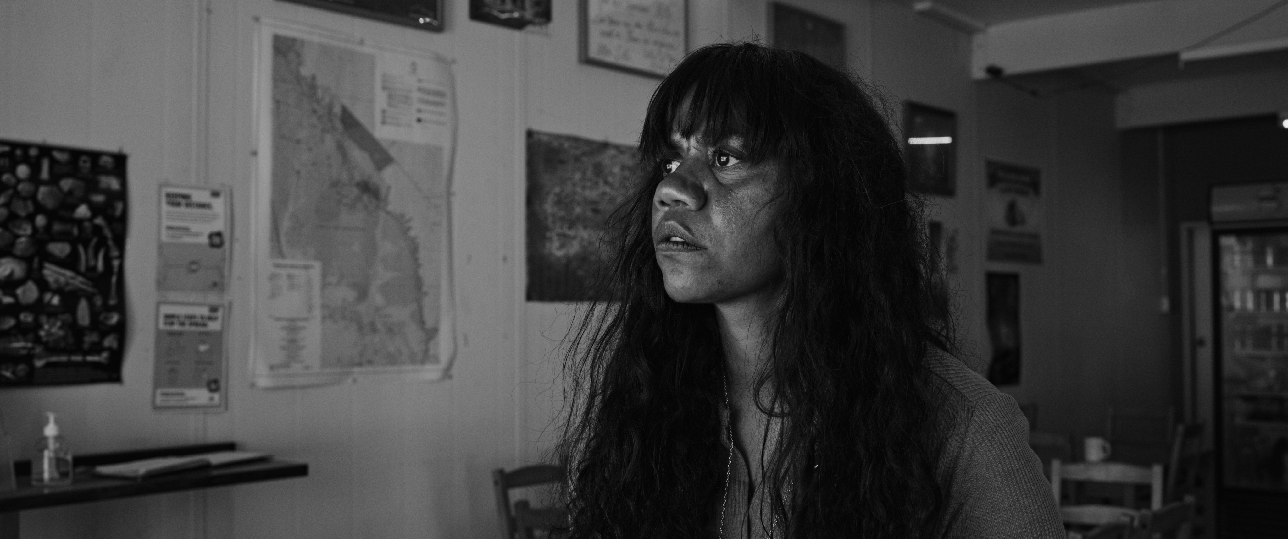 A black and white photo of an Indigenous Australian woman looking off camera. Maps hang on the walls in the room behind her.