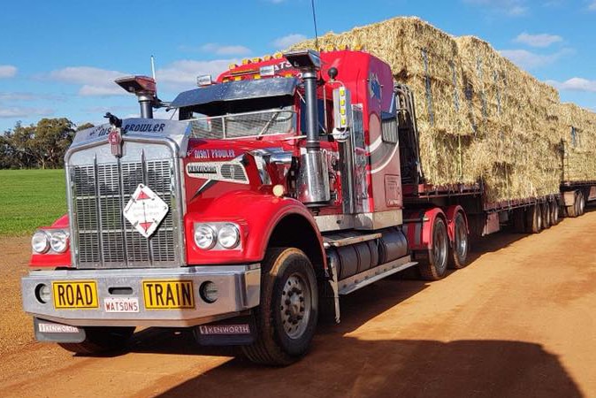 A red road train laden down with hay in bright sunshine.