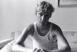 Black and white image of Troye Sivan. He is wearing a white tank top and sitting on a couch.