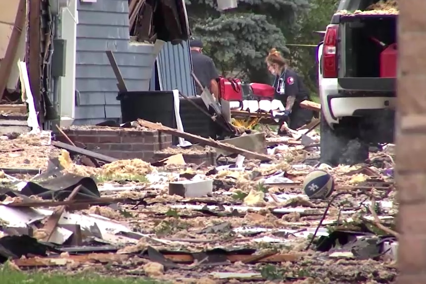 Debris lies scattered across a front lawn in front of a damaged house as emergency personnel wheel a gurney into the house.