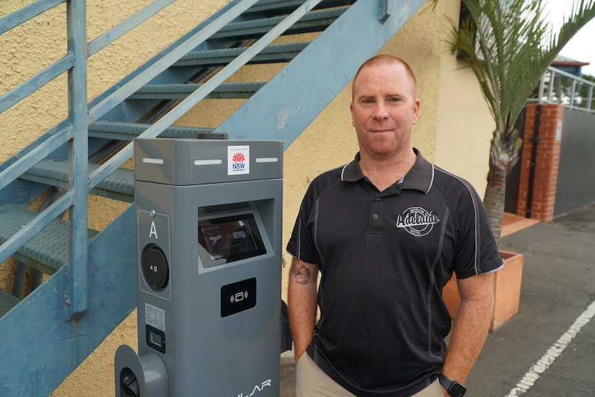 A man looks at the camera while standing next to electric vehicle chargers at his pub. 