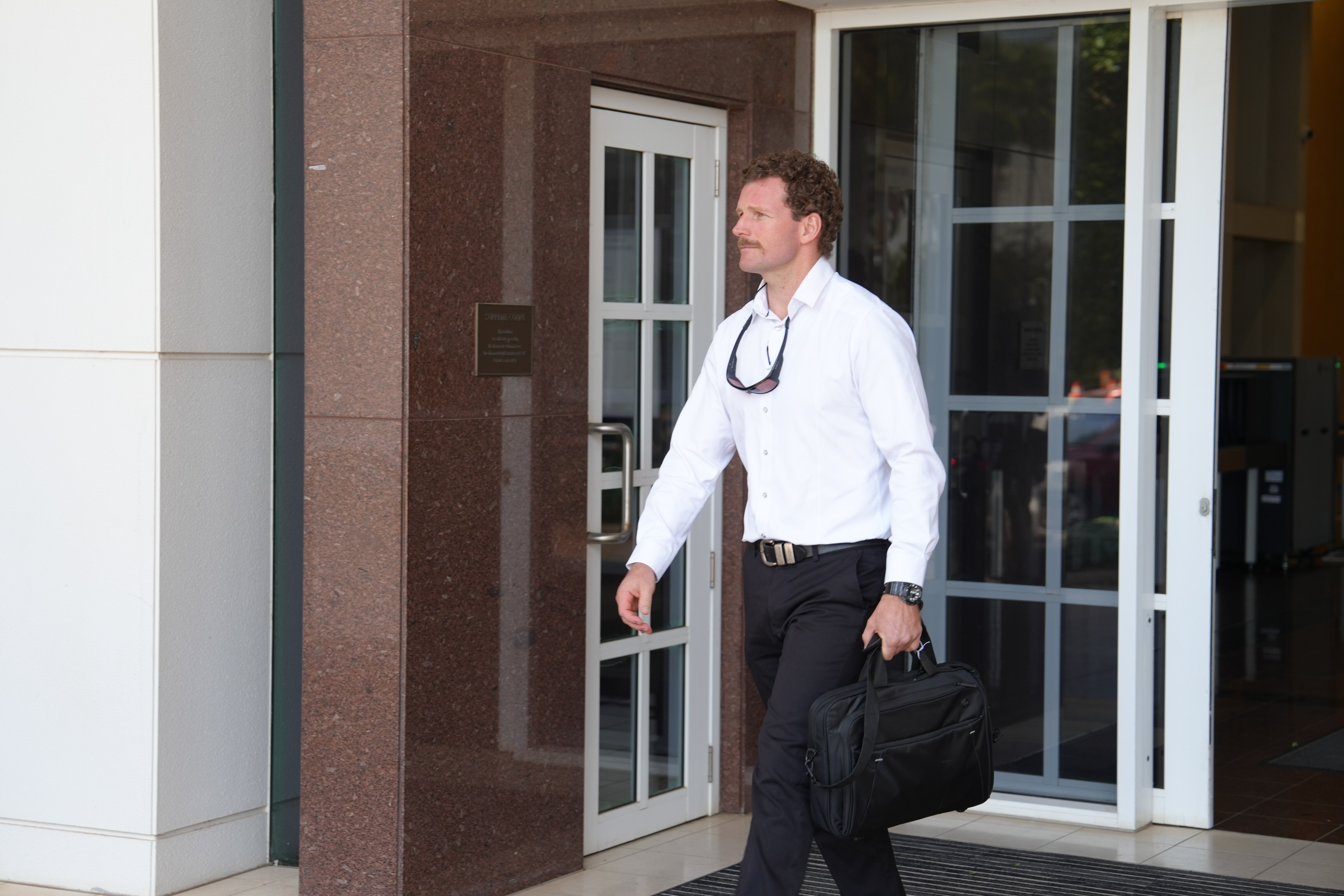 Un hombre de pelo rizado y bigote, vestido con camisa abotonada y portando un maletín, sale de la sala.