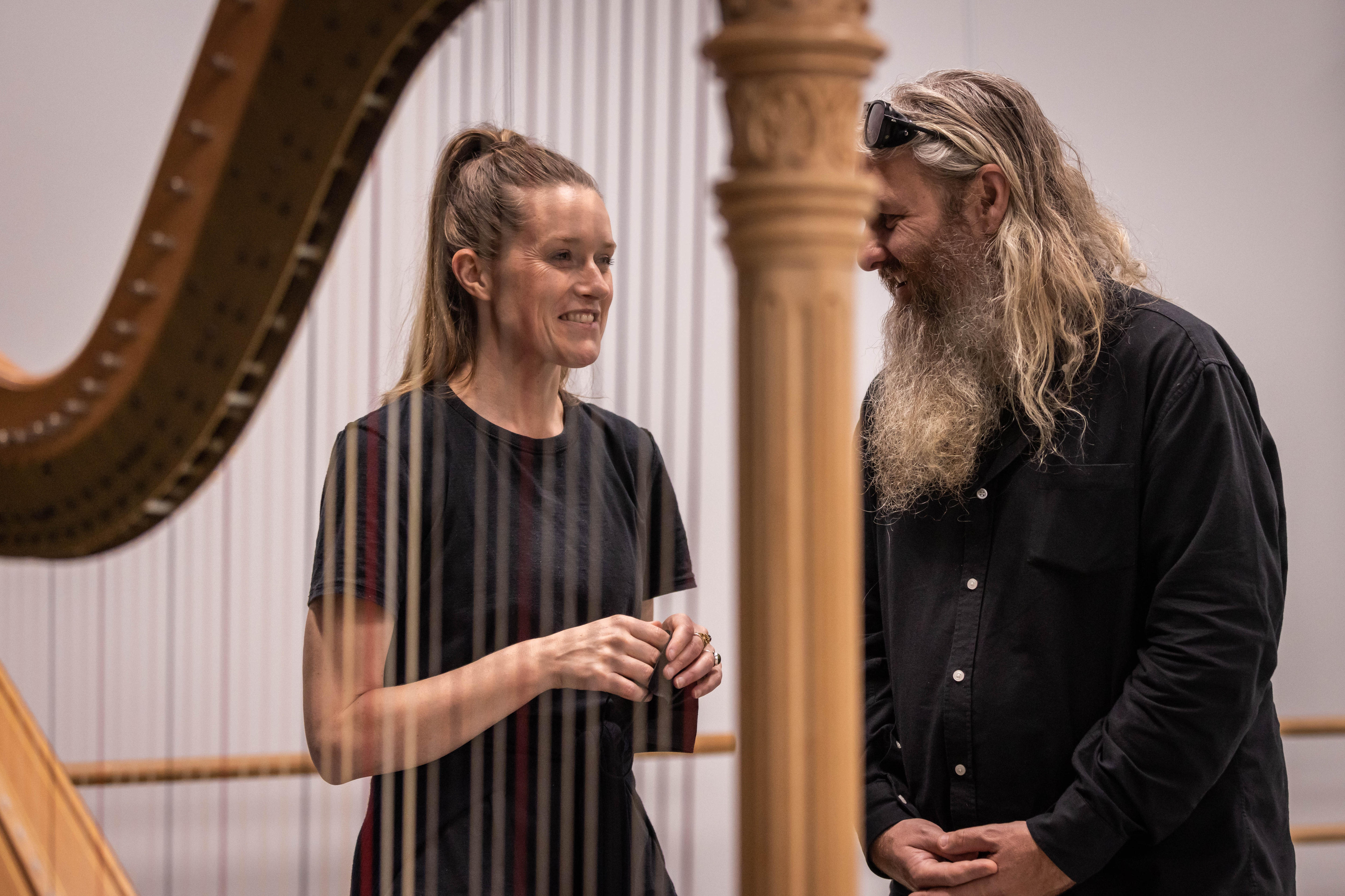 A blonde white woman wears a black t-shirt and speaks to grey long-haired and bearded man in black shirt beside a harp.