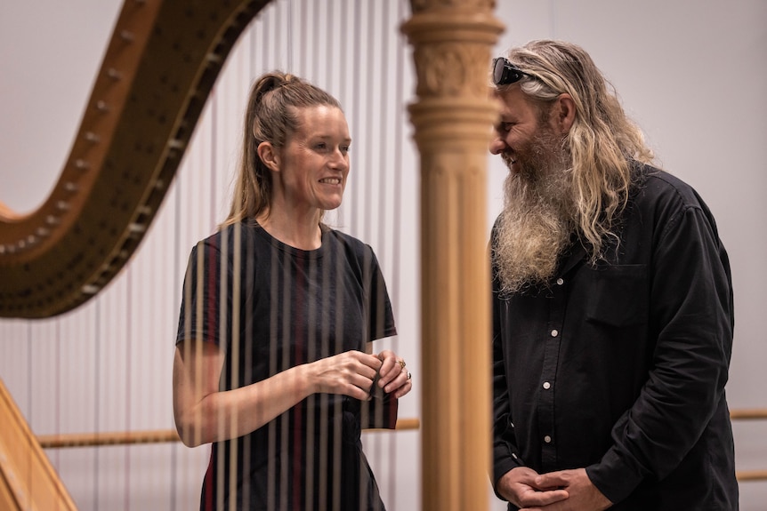 A blonde white woman wears a black t-shirt and speaks to grey long-haired and bearded man in black shirt beside a harp.