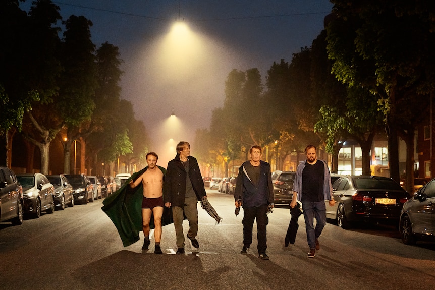 Night-time street-lamp-lit street with the four men walking in a row towards camera, one wearing underpants, shoes and blanket.