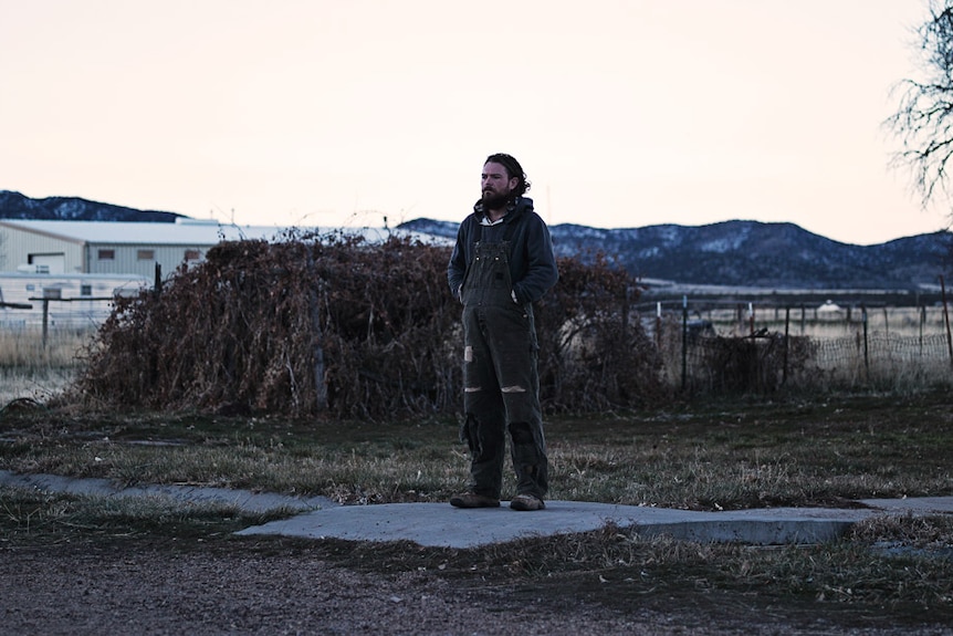 A man with beard and unruly hair stands in a desolate rural area