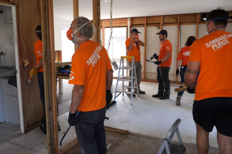 Six people working in orange Samaritan's t-shirts in a home stripped of interior wall sheeting bearing the wooden frame.