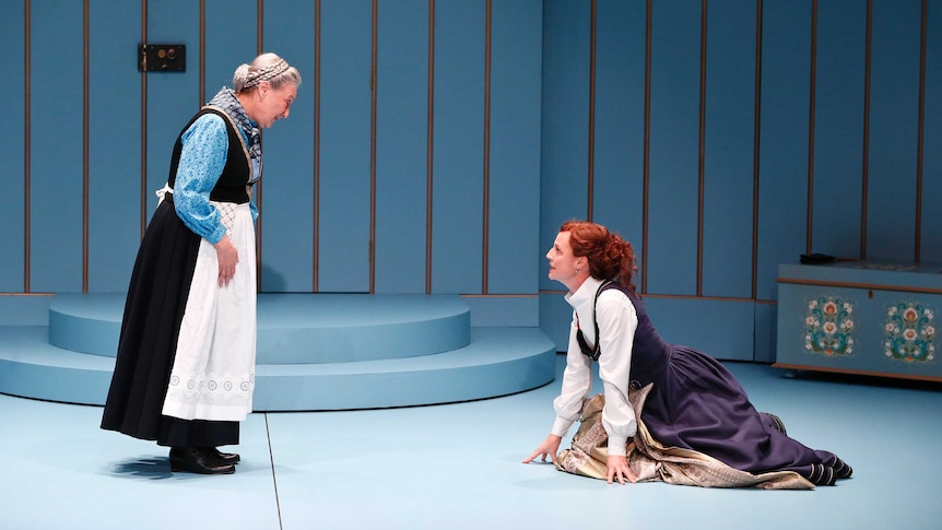 On stage in front of a blue wall, a woman in an apron stands over another woman crouched on the floor.