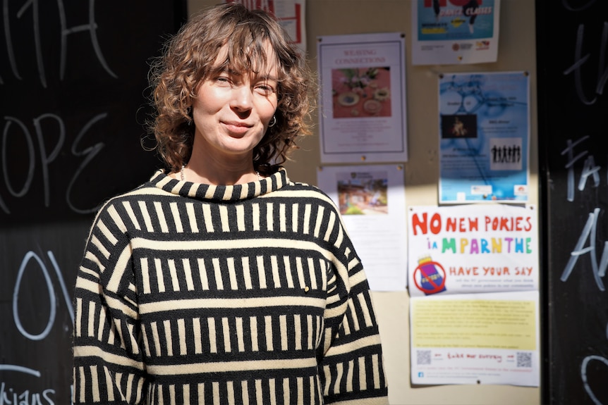 A smiling young woman, short curly brown hair, stands in front of an anti-pokies poster, sun in her face, sun in her face.